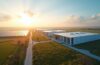 Ein strahlendes, editorial aufgenommenes Foto zeigt einen hochmodernen, modularen Rechenzentrums-Campus unter natürlich warmem Sonnenlicht, eingebettet in eine weitläufige, nachhaltige Landschaft mit Solarfeldern im Vordergrund und klar blauem Himmel, das eine zukunftsweisende digitale Infrastruktur symbolisiert und Optimismus für den globalen Ausbau energieeffizienter Großprojekte ausstrahlt.