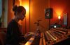 Ein warm erleuchtetes Tonstudio mit einer jungen Musikerin, die konzentriert am Mischpult arbeitet, umgeben von modernen Instrumenten und subtilen KI-Symbolen in Form abstrakter Lichtreflexionen, die die spannende Verbindung von menschlicher Kreativität und künstlicher Intelligenz in der Musikbranche widerspiegeln.