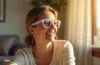 Eine junge Frau sitzt in einem hellen, modern eingerichteten Raum, trägt eine stylische, dezente Brille mit klarem, leicht futuristischem Design und blickt lächelnd konzentriert geradeaus, während sanftes Tageslicht ihre Gesichtszüge warm und freundlich betont – ein Symbol für intelligente Brillentechnologie, die sich dynamisch an ihr Blickverhalten anpasst.