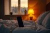 Ein warm beleuchteter Nachttisch im heimischen Schlafzimmer, auf dem ein modernes Smartphone neben einer sanft glimmenden Lampe liegt, während zarte Morgen- oder Abendsonne durch ein Fenster fällt und so die unsichtbaren, digitalen Verbindungen des Geräts subtil spürbar macht – ein ehrlicher und zugleich beruhigender Moment voller technischer Präsenz im Alltag.